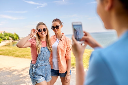 leisure and friendship concept - happy smiling teenage girls or best friends in sunglasses being photographed by smartphone at seaside in summer