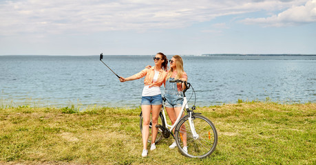 leisure, technology and friendship concept - happy smiling teenage girls or friends with bicycle taking picture by selfie stick on smartphone at seaside in summer