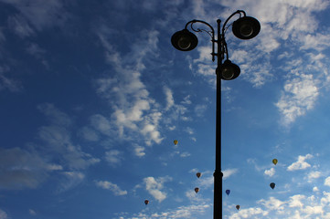Street lamp pole with blue sky background and hot air balloons seen flying in the distance.