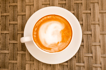 Cappuccino in white cup on cane wood table at cafe. Top view. Copyspace. Bewerages, coffee lovers and morning menu concept. Stylish toning.