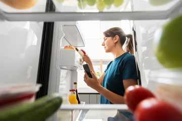 healthy eating and diet concept - woman at fridge with smartphone making list of necessary food at home kitchen
