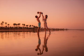 Fototapeta premium A young family travels the world. Parents throw in the arms of a child at sunset. Reflection in water.