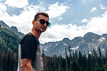 Young attractive man in black sunglasses and casual t-shirt looking at the camera, beautiful mountainss on the background, copyspace for your text