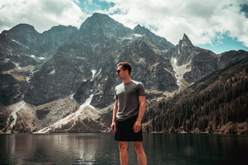 Young man in black sunglasses and casual clothes standing in front of mountain lake and mountain range