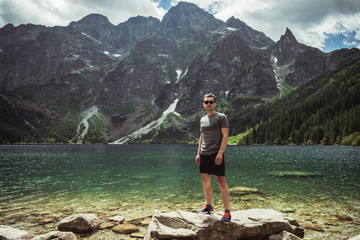 Young man in black sunglasses and casual clothes standing on the big stone in front of the mountain lake and range