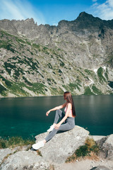 Naklejka na ściany i meble Young beautiful sporty girl in grey pants sitting on the big rock and posing on the background of mountain lake, active lifestyle concept