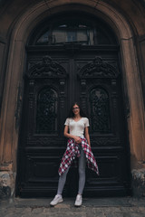 Full length portrait of beautiful young brunette girl in casual clothes and glasses that standing in front of the big old antique black door