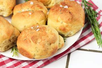 homemade fresh herb bread rolls of Provencal herbs with salt and a sprig branch of rosemary. Bread rolls bakery style on white plate and red and white towel in a cage italian style