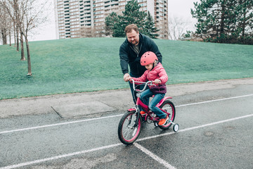 Caucasian father dad training helping girl daughter to ride bicycle. Preschooler child kid in pink helmet with bike on backyard road outside on spring day. Seasonal child family outdoor activity