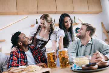 two cheerful waitresses in traditional german costumes serving beer for multicultural friends in pub