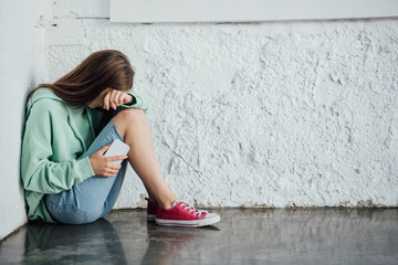 sad crying girl sitting near textured wall and holding smartphone