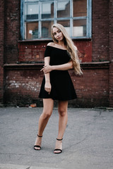 Young blond woman with long hair in a black dress near a brick red wall. Stylish girl in the city.