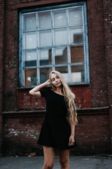Obraz na płótnie Canvas Young blond woman with long hair in a black dress near a brick red wall. Stylish girl in the city.