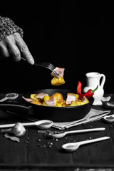 hand holds a fork with fried potatoes and bacon meat over a pan on a dark background