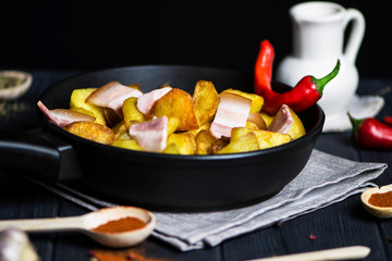 spicy fried potatoes with bacon in a pan and various spices on a dark background