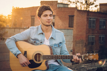 Young man playing guitar in city on sun rays background. Handsome hippie style guitarist engrossed...