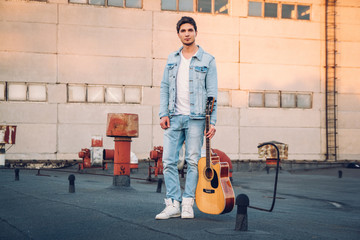Young man holding guitar in city on sun rays background. Handsome hippie style guitarist engrossed...