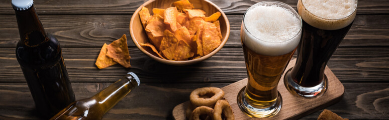 panoramic shot of glasses of light and bark beer near bottles and snacks on wooden table