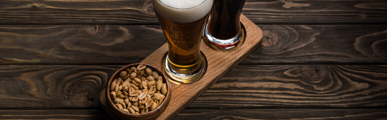 panoramic shot of glasses of dark and light beer near bowl with roasted peanuts on wooden table