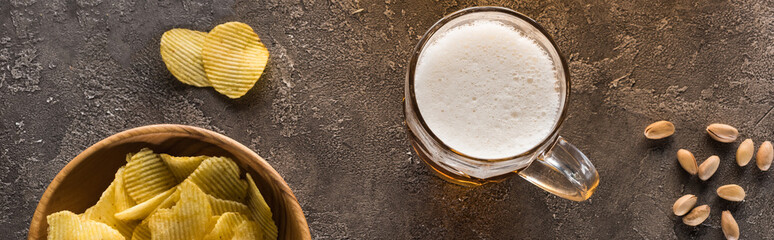 panoramic shot of mug of light beer near crisps and scattered pistachios on brown textured surface