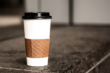 White Coffee Cup With Black Lid and Insulation Sitting On Table With Napkin In Morning For Breakfast