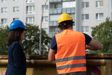 Back turned construction manager wearing safety jacket and helmet checking projects discussing with a female engineer