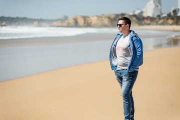 Young man enjoying sunny days on the beach. Portrait beautiful guyl walking and playing with sand. Smiling male posing near ocean. Vacation in Portugal