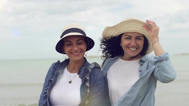 Happy joyful young women best friends on vacation with straw hats looking at the ocean and then looking at camera and smiling.