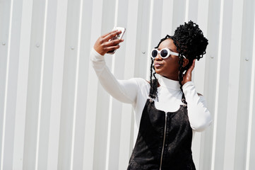 African woman in denim overalls skirt, white sunglasses posed against white steel wall and making selfie by phone.