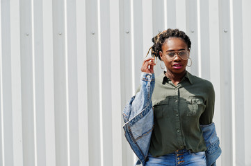 African woman with dreads hair, in jeans jacket and shorts, wear eyeglasses posed against white steel wall.