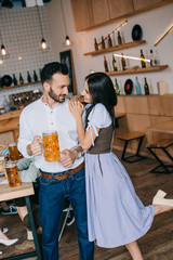 smiling young man holding glass of beer while standing near beautiful woman in traditional german costume