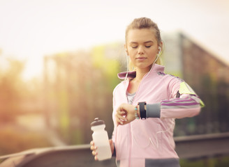 Woman running at sunset in city park using smart watch and earbuds. Athlete listening to music during workout at park and checking her results on a tracker