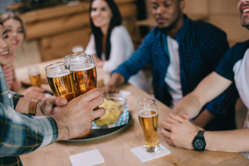 cropped view of man holding glasses of light beer near multicultural friends in pub