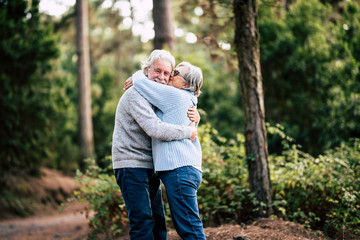 Couple of happy and cheerful people caucasian senior hug with love and smile with defocused green forest in background - outdoor leisure activity for retired matures- happiness for old