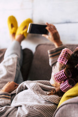 Relaxed young caucasian woman at home use smartphone lay down onthe sofa with home dress and curlers for hair- people and beauty carefree concept