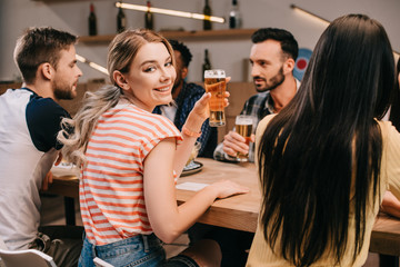 selective focus of cheerful young woman smiling at camera while holding glass of light beer