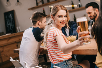 selective focus of smiling young woman looking at camera while holding glass of light beer