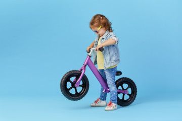Fototapeta na wymiar Little girl learns to keep balance while riding a bicycle. Stylish child in sunglasses isolated on blue background