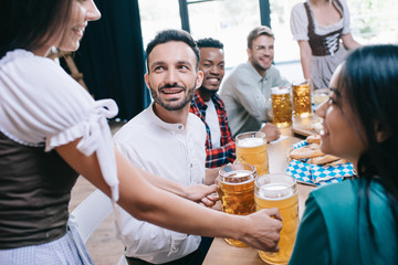 cropped view of waitresses in traditional german costumes serving beer for multicultural friends in pub