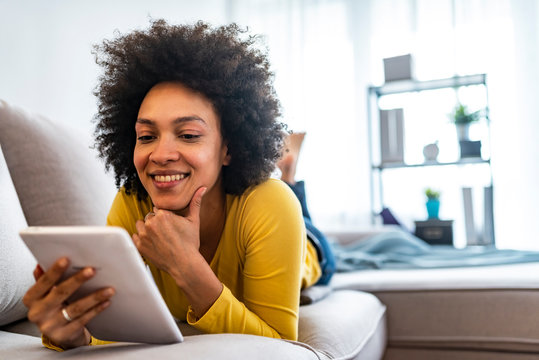 Portrait Of Young Happy African Woman Using Digital Tablet. Chilling freelancer on the beige couch at home doing online shopping, so comfortable, in fashioanble outfit, denim pants