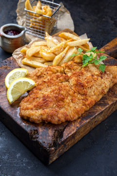 Frittiertes Wiener Schnitzel von der Oberschale vom Kalb mit Pommes Frites und Zitrone als closeup auf einem alten rustikalen Schneidebrett