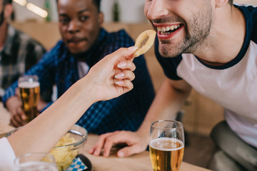 cropped view of woman feeding cheerful man with fried onion ring while sitting in pub with friends