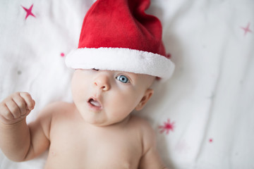 Surprised baby in red Santa hat laying on a bed with red stars with opened mouth and arm up