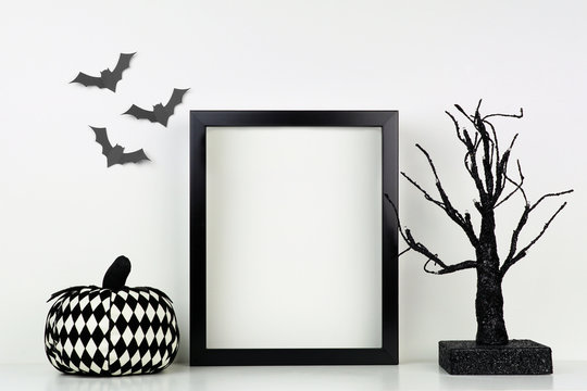 Mock up black frame with pumpkin and spooky tree decor on a shelf or desk. Halloween concept. Portrait frame against a white wall with bats.