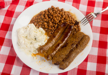 Spicy Juices from Sweet and Spicy Sausages on Plate with Potato Salad and Beans against Red Plaid BG