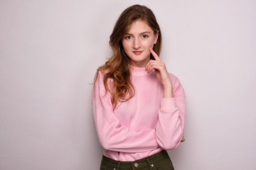 A girl in a pink sweater stands on a white background, looks at the camera, propping her cheek with...