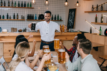 cheerful young man holding mugs of light and dark beer in pub near multicultural friends