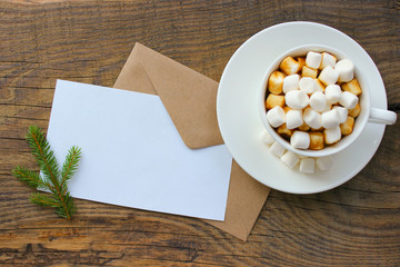 Cup with homemade Christmas hot chocolate drink, marshmallows and holiday greeting card on wooden background.