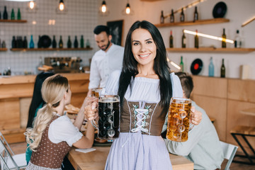 beautiful waitress in traditional german costume holding mugs of light and dark beer and smiling at camera