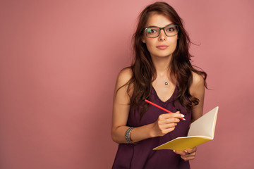 A dark-haired girl in a purple top and glasses stands on a pink background with a yellow notebook and pencil, looks at the camera